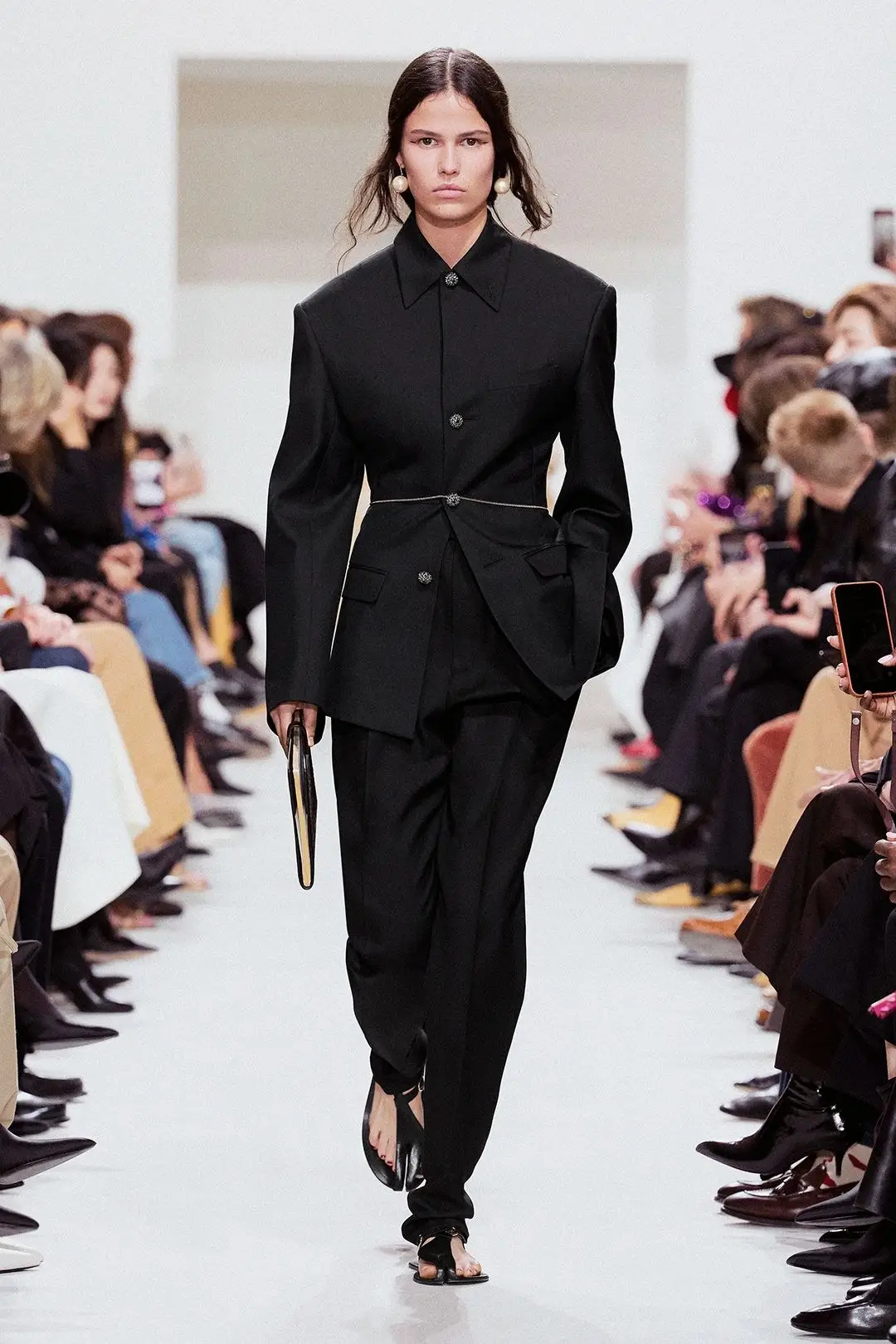 Celine Spring 2027, look 2. A black tailored jacket and trousers on a white runway between rows of guests.