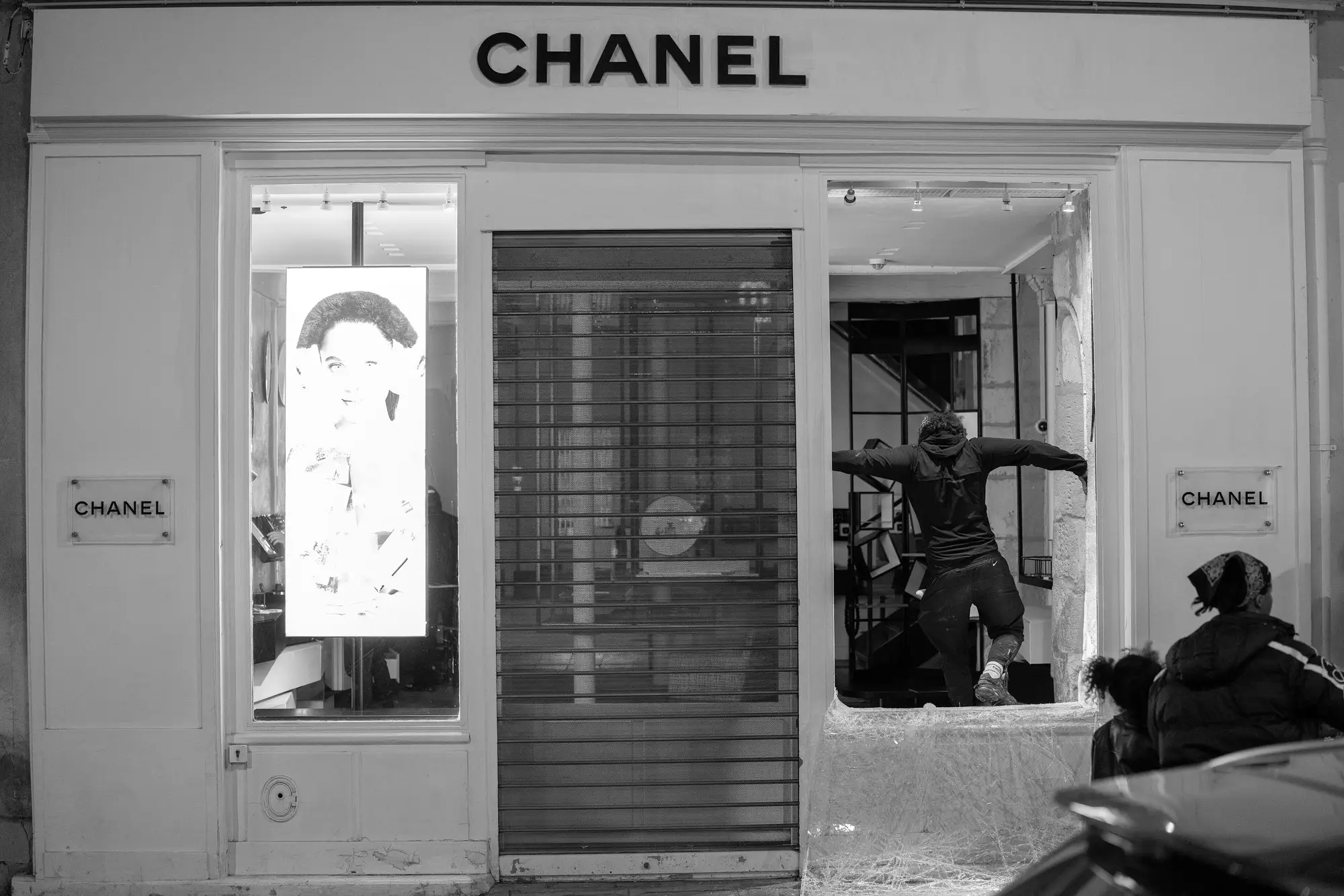 Night in Paris, 17 October 2026. A Chanel boutique with its shutter half down and a smashed window, a person climbing through the gap.