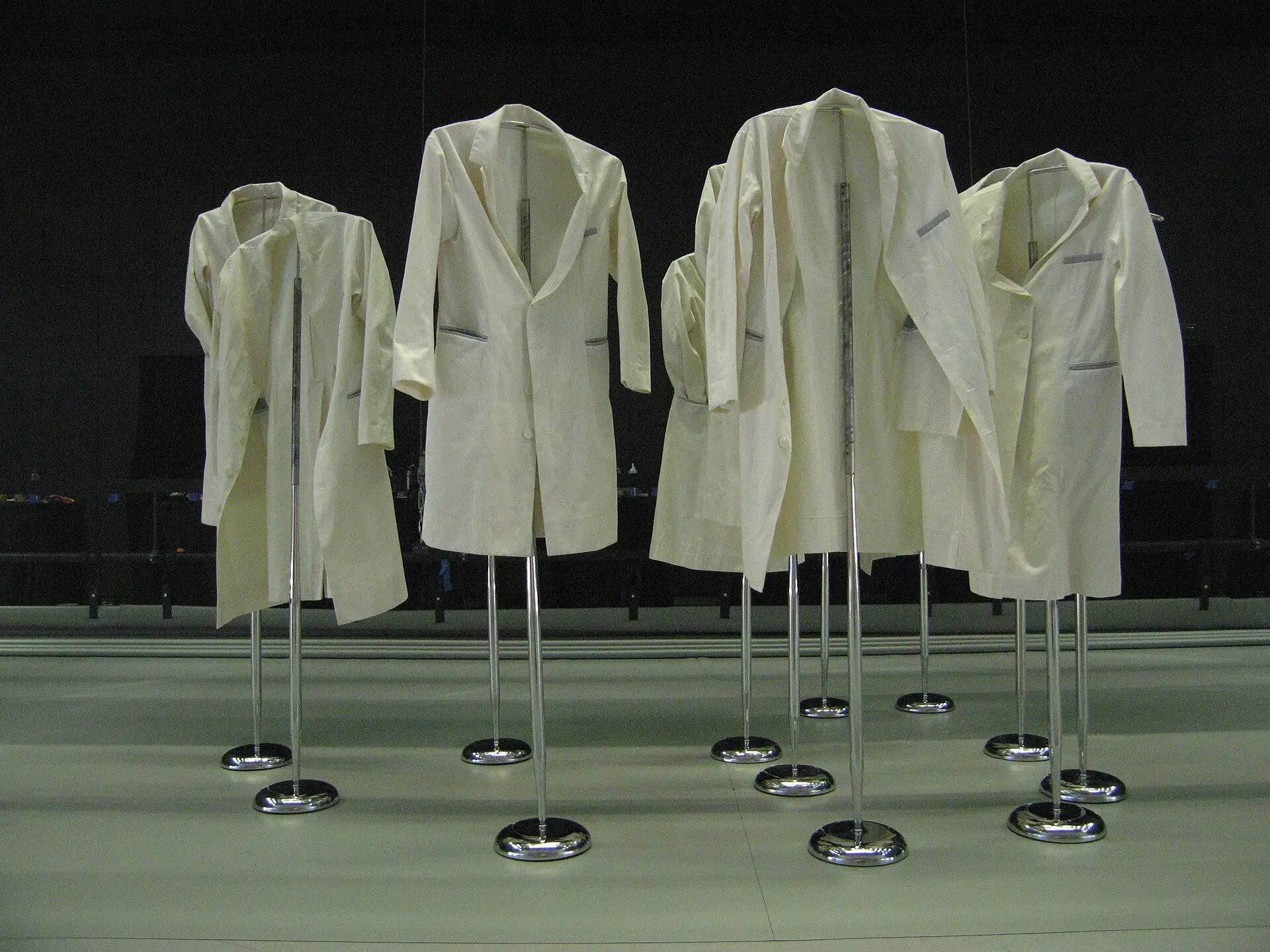 Five white lab coats hung on stands in a row, like empty people.