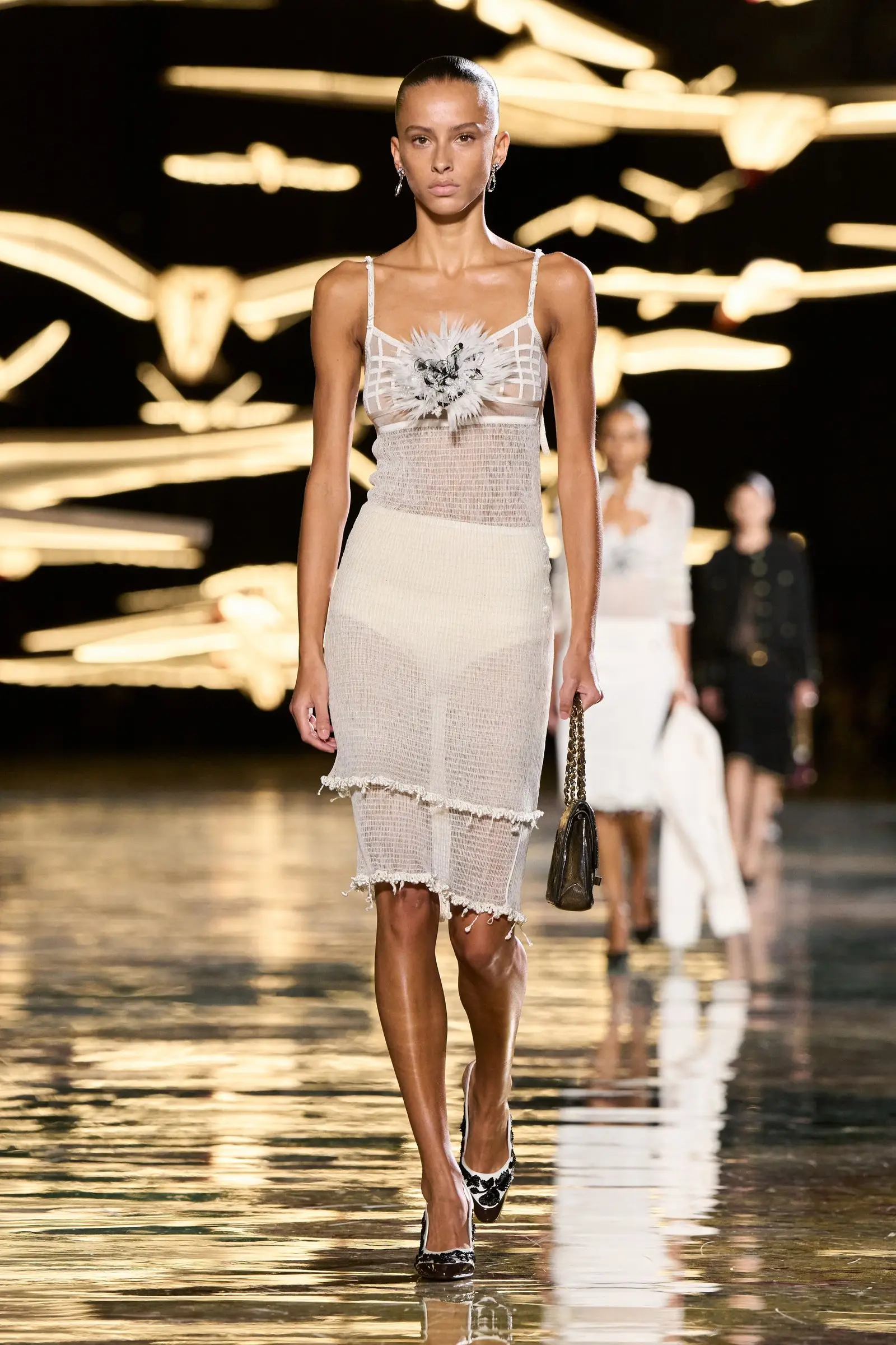 Chanel SS27, Look 1. The model Dyanira Hurbuscher in white, carrying a black quilted bag on a gold chain in her left hand.