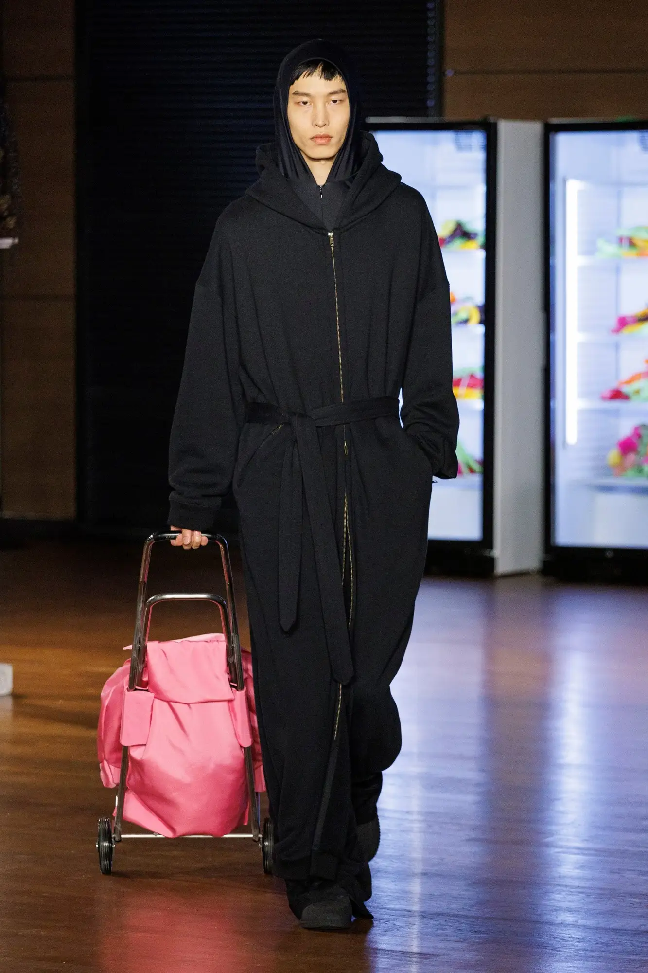 Look 20. Black hooded coat, a pink trolley.