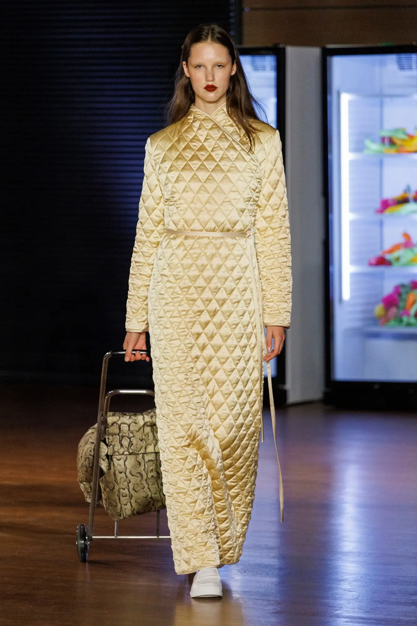 Marie Adam-Leenaerdt SS27, Look 38. A long, shiny quilted dress in pale gold, worn with a wheeled shopping trolley in snake print, in front of lit fridges full of fruit.