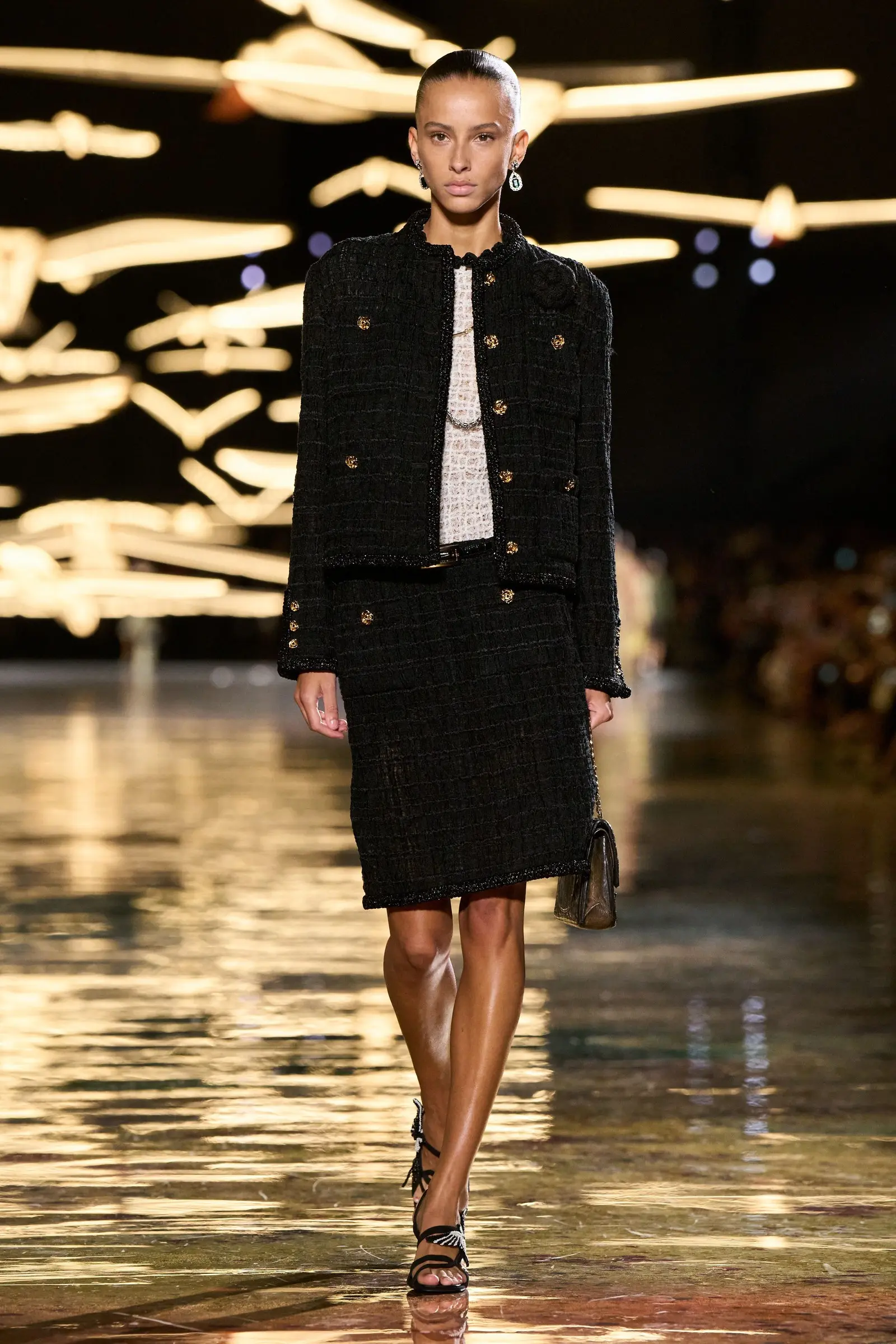Chanel SS27, Look 83. A model in a black smocked tweed jacket and skirt, a black quilted bag in her right hand.