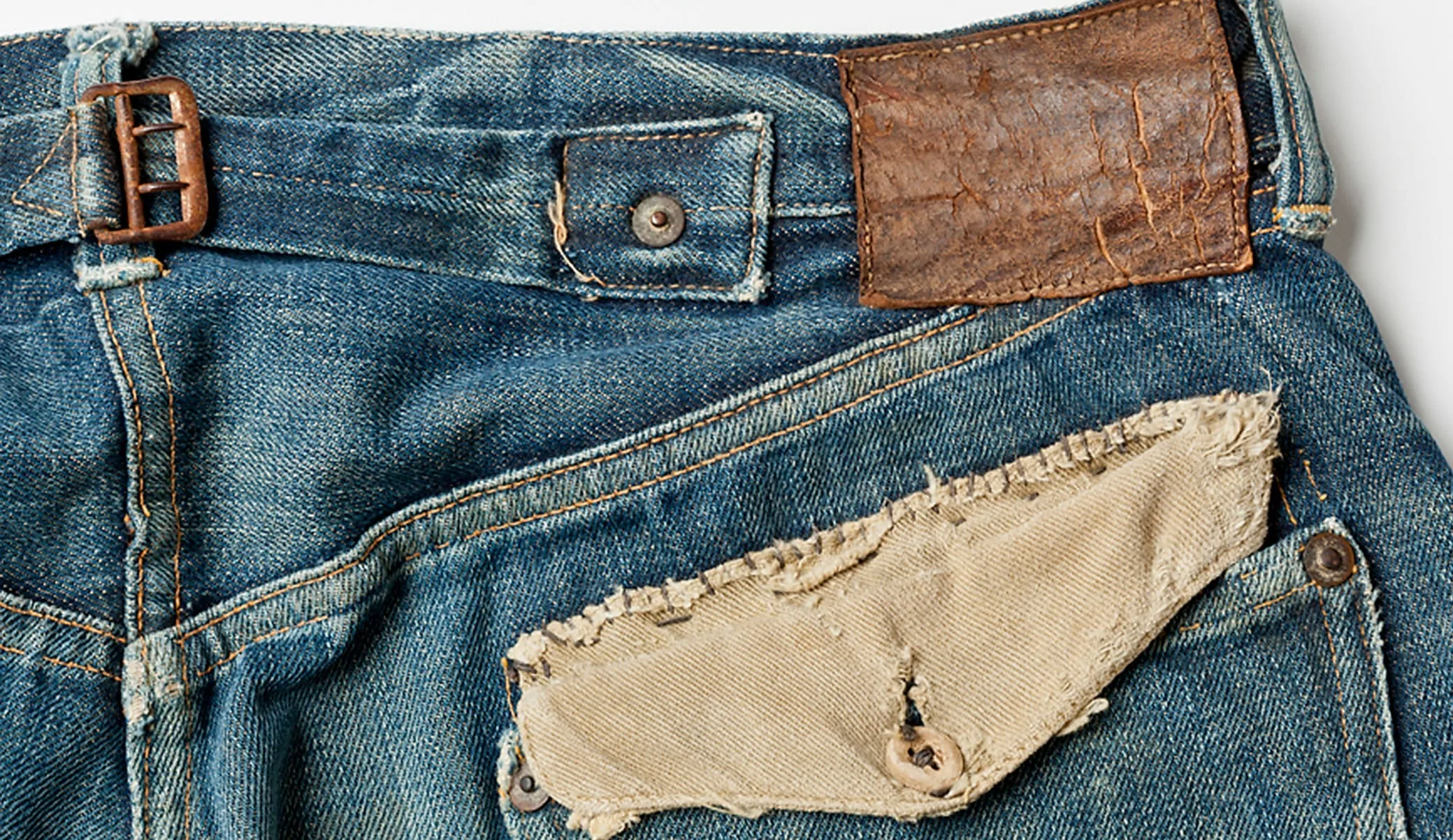 Worn Levi's jeans up close: faded blue denim, a cracked leather patch, a worn patch over the back pocket and a metal back buckle.