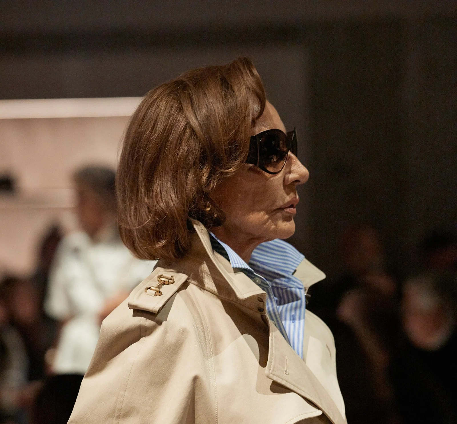 Closer: Naty Abascal in the trench, collar up over a blue striped shirt, epaulettes on the shoulders, sunglasses on.